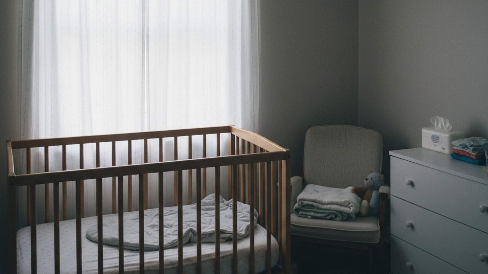 Chambre de bébé avec un berceau en bois légèrement usé près d'une fenêtre, rideau translucide, lumière douce de fin de matinée, quelques vêtements posés sur le dossier d'une chaise